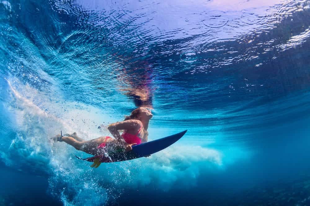 Young active girl wearing bikini in action - surfer with surf board dive underwater under big ocean wave. Family lifestyle people water sport adventure camp and beach extreme swim on summer vacation.
