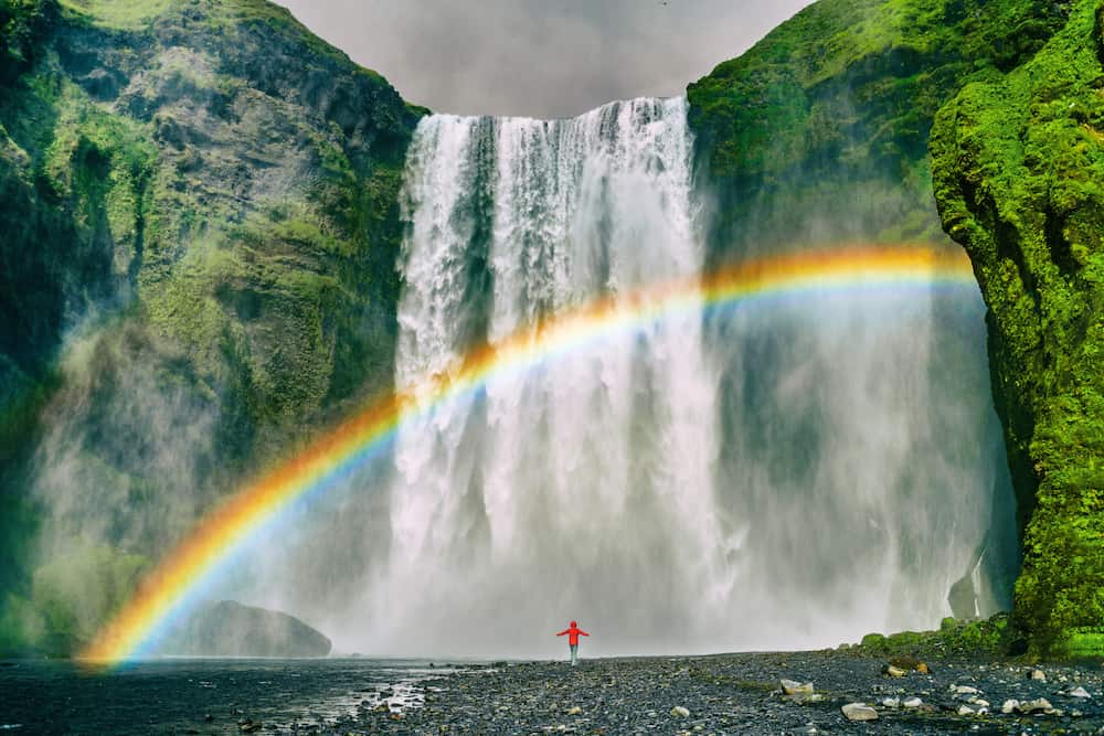 Iceland waterfall travel nature famous tourist destination. Skogafoss waterfall with rainbow and woman under water fall in magical landscape popular Europe attraction.
