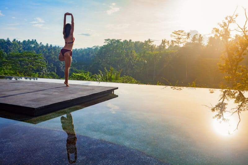 Good morning with yoga meditating on sunrise background. Active woman in bikini practicing on villa poolside to keep fit and health. Woman fitness training sport activity on summer family holiday.