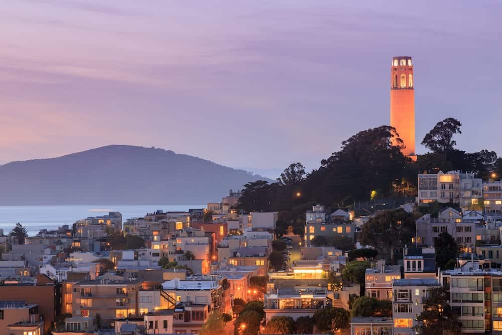 Coit Tower on Telegraph Hill with San Francisco Bay and Angel Island in the background at dusk. Taken from a downtown building rooftop. San Francisco, California, USA.