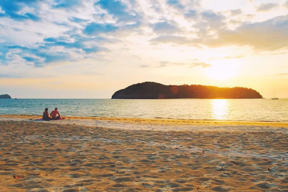 Pantai Tengah Beach at colorful sunset, Langkawi Island, Malaysia. Beach sunset is a golden sunset sky with a wave rolling to shore as the sun sets over the horizon. People relaxing on paradise beach