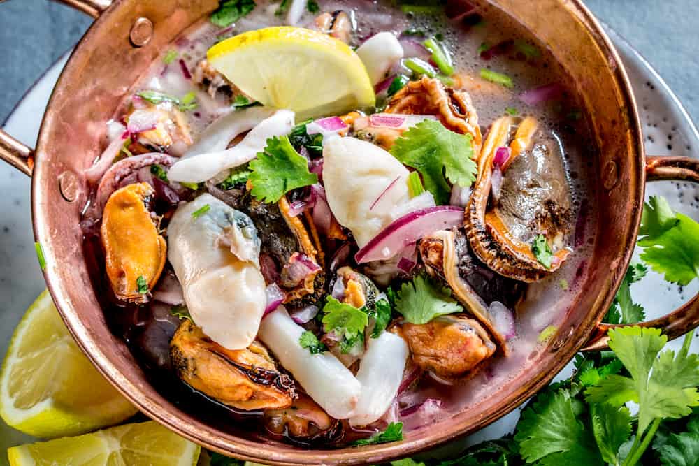 Peruvian Latin American seafood shellfish ceviche cebiche. Raw seafoods - mussels shrimps clams squides marinated in lemon juice with red onion and coriander in cooper bowl gray slate background.