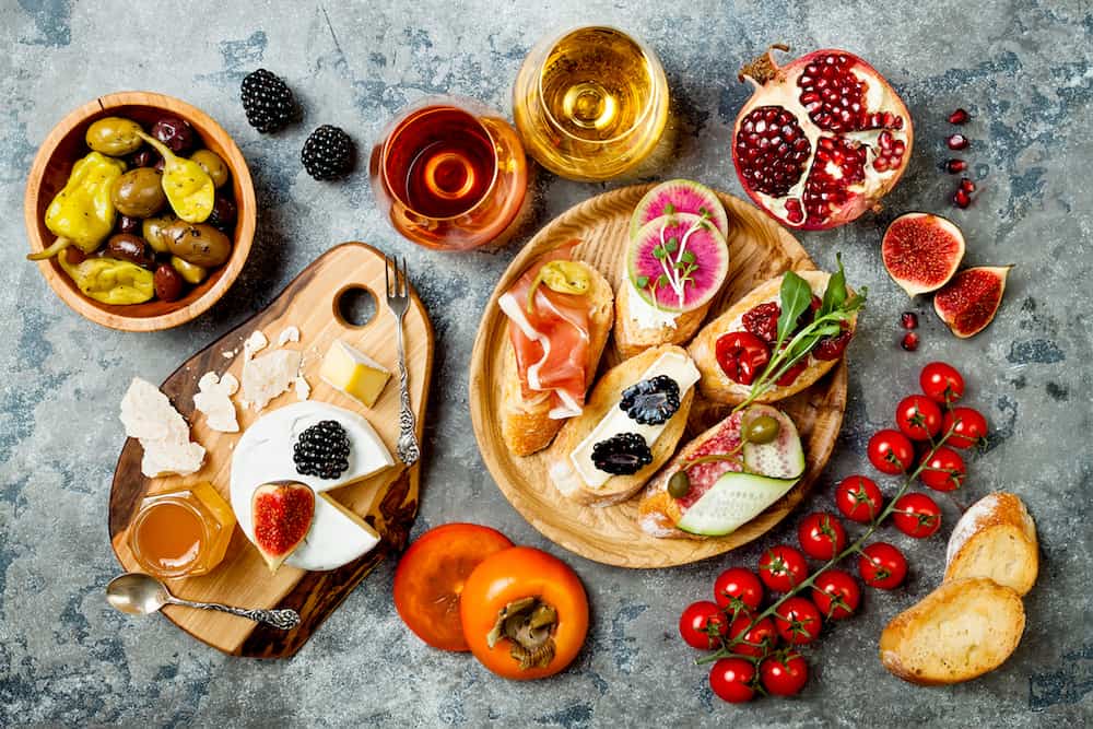 Appetizers table with italian antipasti snacks and wine in glasses. Brushetta or authentic traditional spanish tapas set cheese variety board over grey concrete background. Top view flat lay