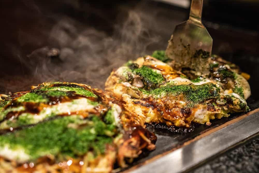 chef is cooking Okonomiyaki on the iron plate, he is using the spatula to put the food on to the plate which is already done