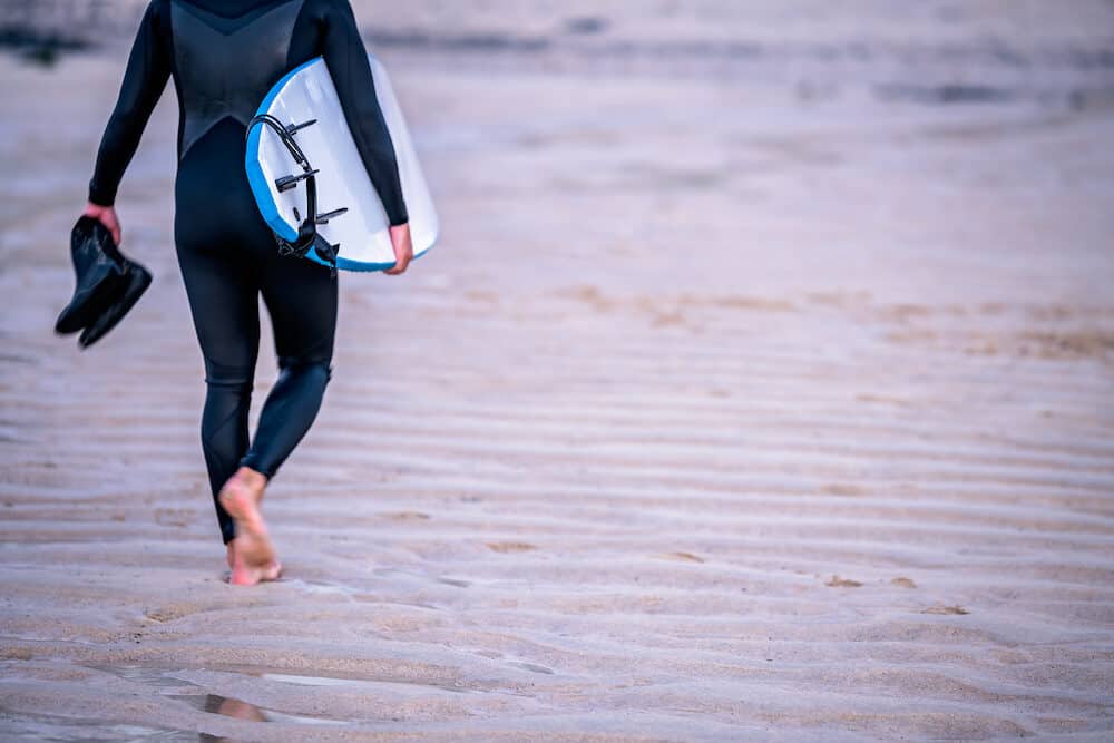 Surfing instructor carrying his shoes and surfing board on the beach in Saint Ives, Cornwall, UK