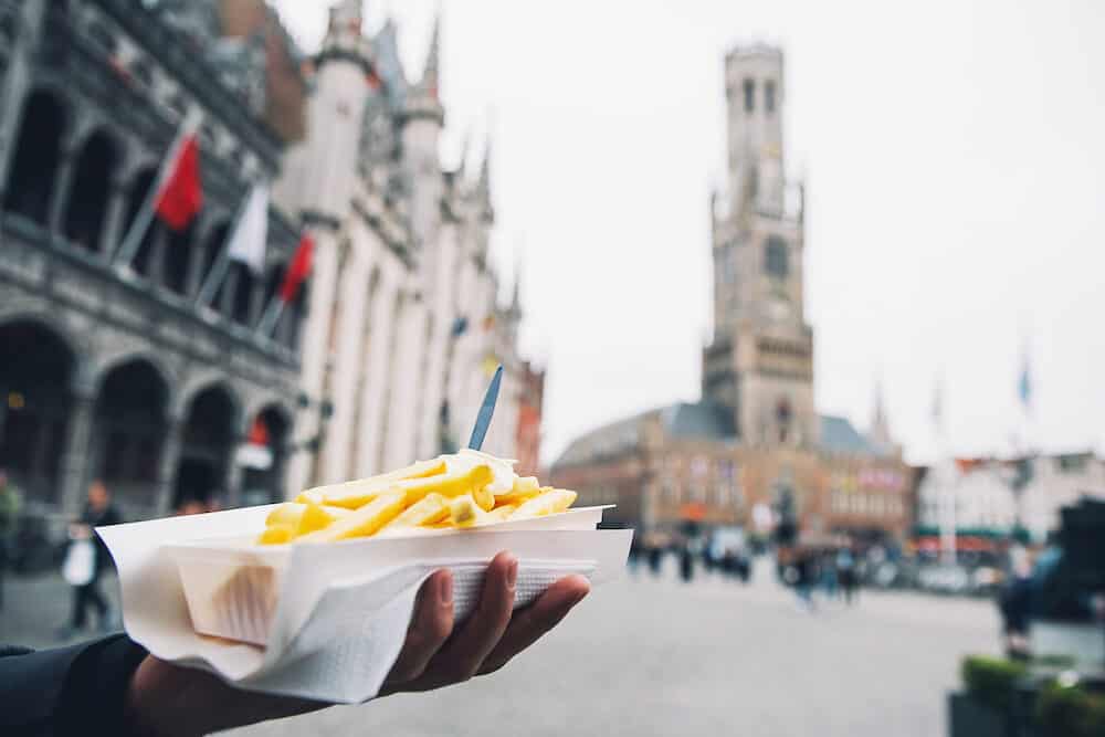 Tourist holds in hand popular street junk food - French Fries with mayonnaise on the background of city tourist streets of Bruges Belgium.