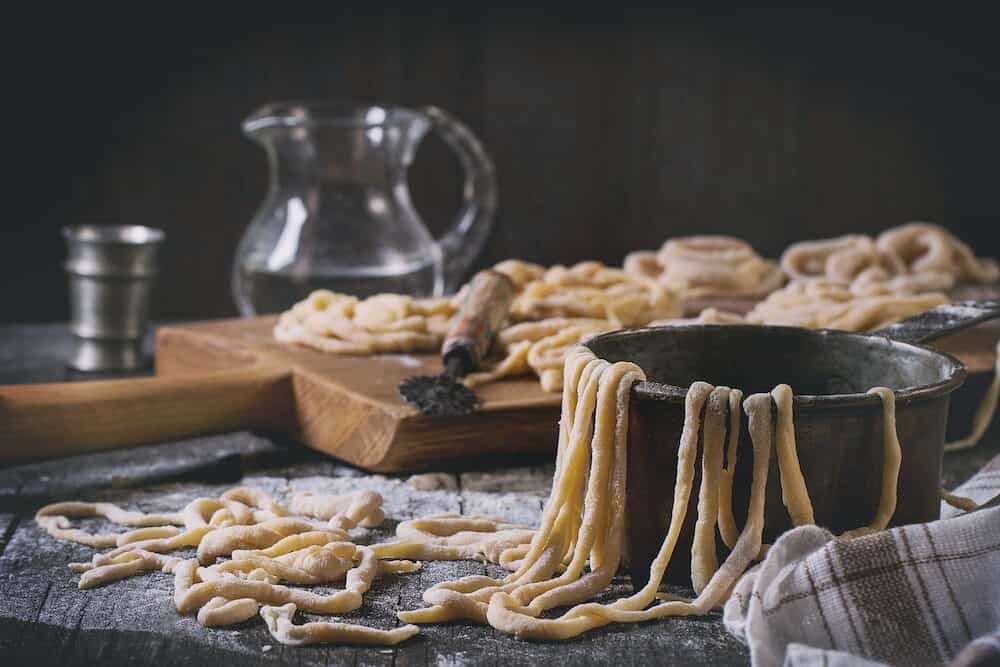 Fresh homemade pici pasta on wood chopping board over old wooden table with flour copper bowl rolling-pin and galss jug of water. Dark rustic style with retro filter effect. See process series