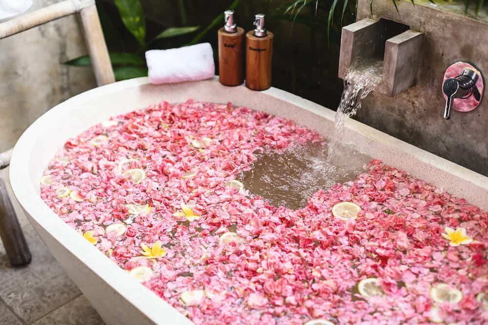 Bath tub with flower petals filling with water. Organic spa relaxation in luxury Bali outdoor bathroom.