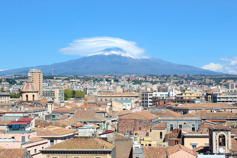 Majestic Mount Etna overlooking the Sicilian city Catania, Italy. Smoke cloud over the famous volcano, snow on the top. Dominant of the cityscape is the cupola of Catania Cathedral of Saint Agatha