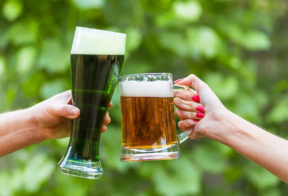 Glass of green Irish beer and mug classic beer in female hands against on a green natural background, Patrick's Day. Mug classic beer in female hands closeup. Glass of green Irish beer closeup.