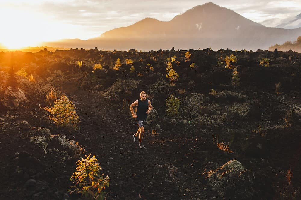 Young male athlete trail running in mountains at sunrise. Amazing black lava volcanic landscape of Bali on background. Adventure sport concept.