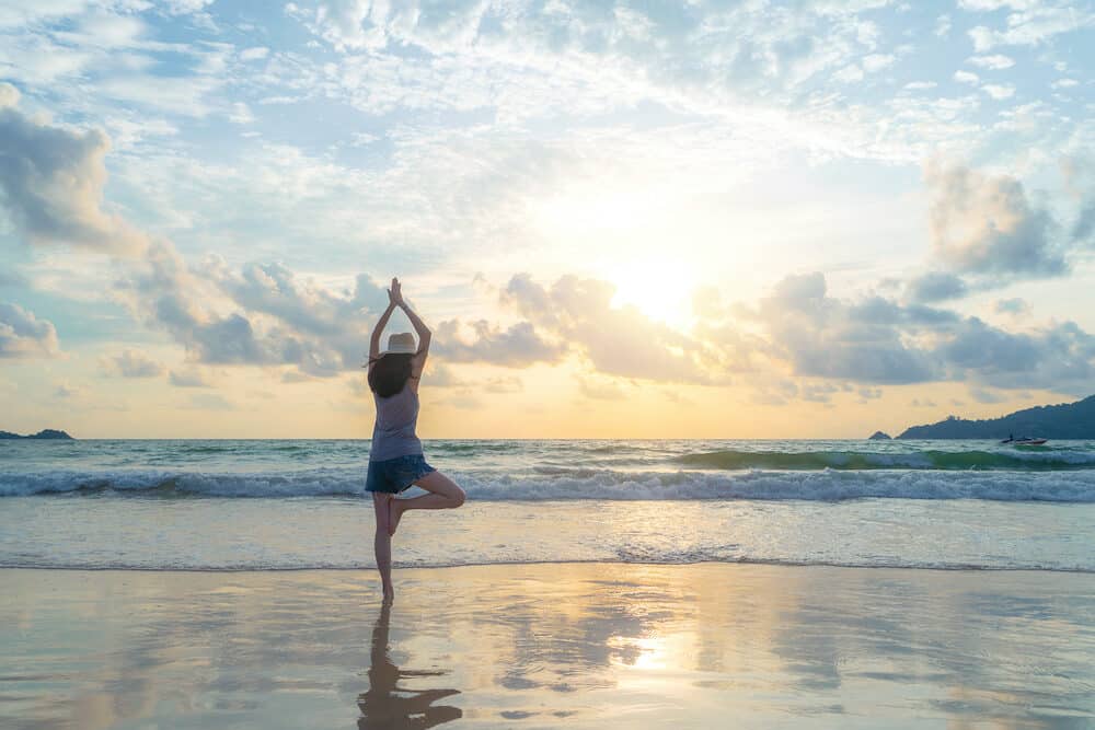 Happy Asian woman practicing yoga or exercising at the beach during travel holidays vacation outdoors at ocean or nature sea at sunset time, Phuket, Thailand