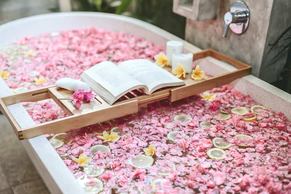 Bath tub with flower petals and lemon slices. Book and candles on a tray. Organic spa relaxation in luxury Bali outdoor bathroom.
