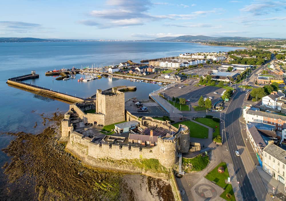 Belfast Lough. Medieval Norman Castle in Carrickfergus in sunrise light. Aerial view with marina, yachts, parking, breakwater, sediments and far view of Belfast in the background