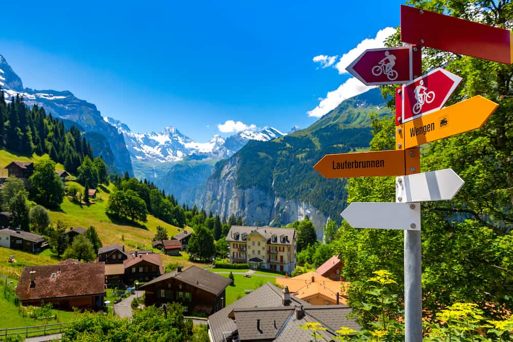 Road sign with arrows for hiking and biking tourist trails pointing fork in the road from village Wengen to Lauterbrunnen, Bernese Oberland, Switzerland. The Jungfrau is visible in the background
