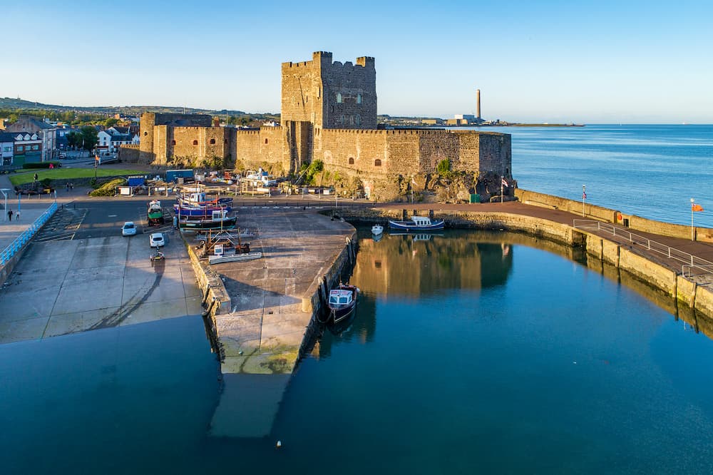 Medieval Norman Castle and harbor with boat ramp and wave breaker in Carrickfergus near Belfast, Northern Ireland, UK. Aerial view in sunset light. Old power plant in the background