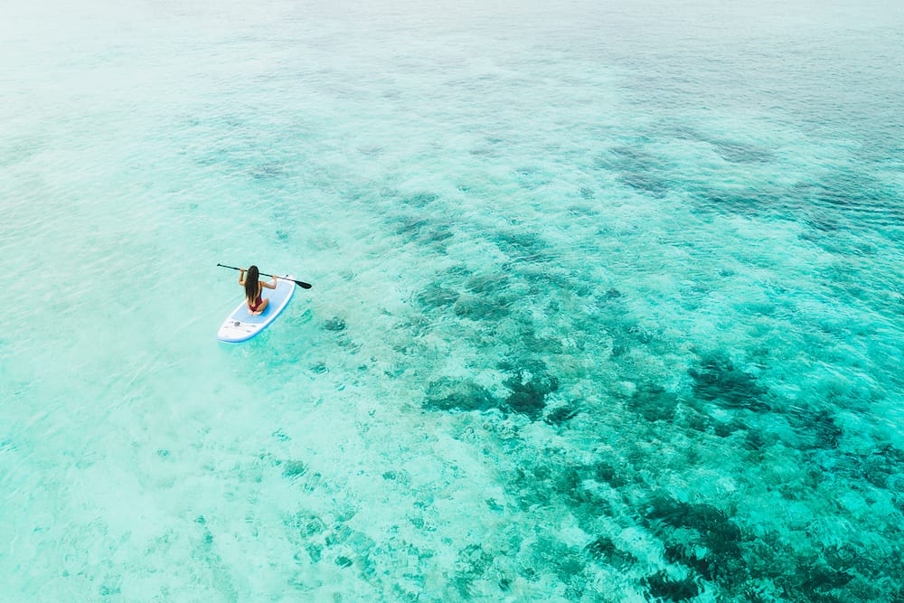 Woman paddling on sup board and enjoying turquoise transparent water and coral reef. Tropical travel, wanderlust and water activity concept. View from back.
