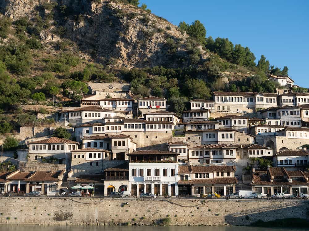 Berat, Albania - Ottoman houses with multiple windows built on a hillside in Berat, Albania with the River Osum and businesses in the foreground.