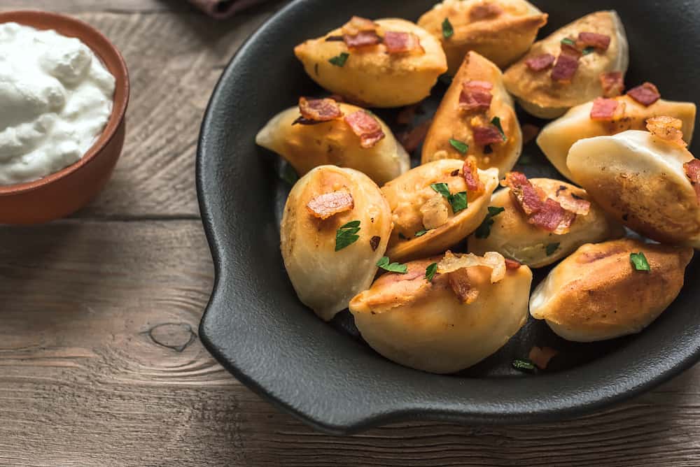 Fried dumplings stuffed with potato and meat (pierogi) sprinkled with bacon and parsley and sour cream on wooden table, top view, copy space.