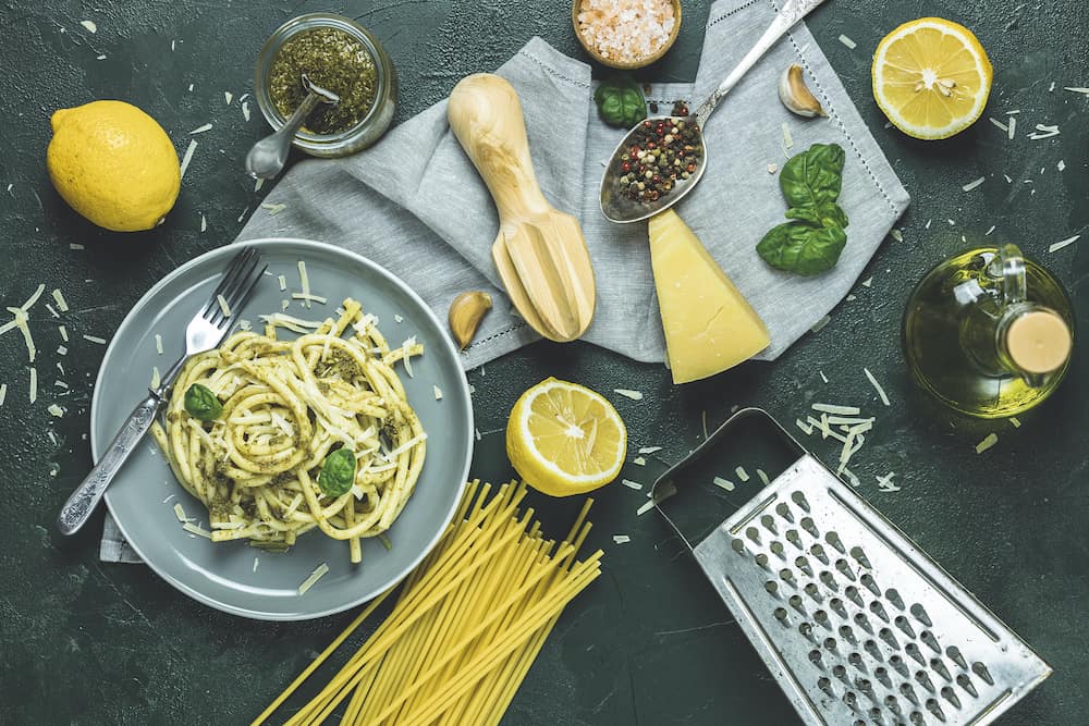 Italian traditional perciatelli pasta by genovese pesto sauce in gray dish served with ingradients on dark green concrete table. Flat lay on dark green surface