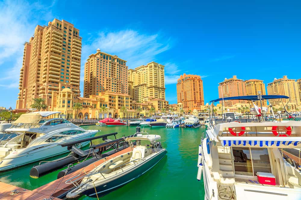 Doha, Qatar - Marina corniche promenade in Porto Arabia at the Pearl-Qatar, Doha, with residential towers and luxury boats and yachts in Persian Gulf, Middle East. Sunny blue sky.