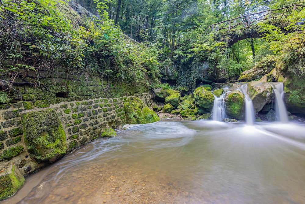 Schiessentümpel waterfall, stone bridge over the Black Ernz river, brick wall with moss, water flowing between the rocks, wild vegetation in the background, Mullerthal Trail, Luxembourg. Long exposure