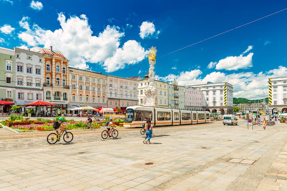 Austria: View of the Main Square in Linz on a sunny summer day. A group of people riding bikes towards the city center