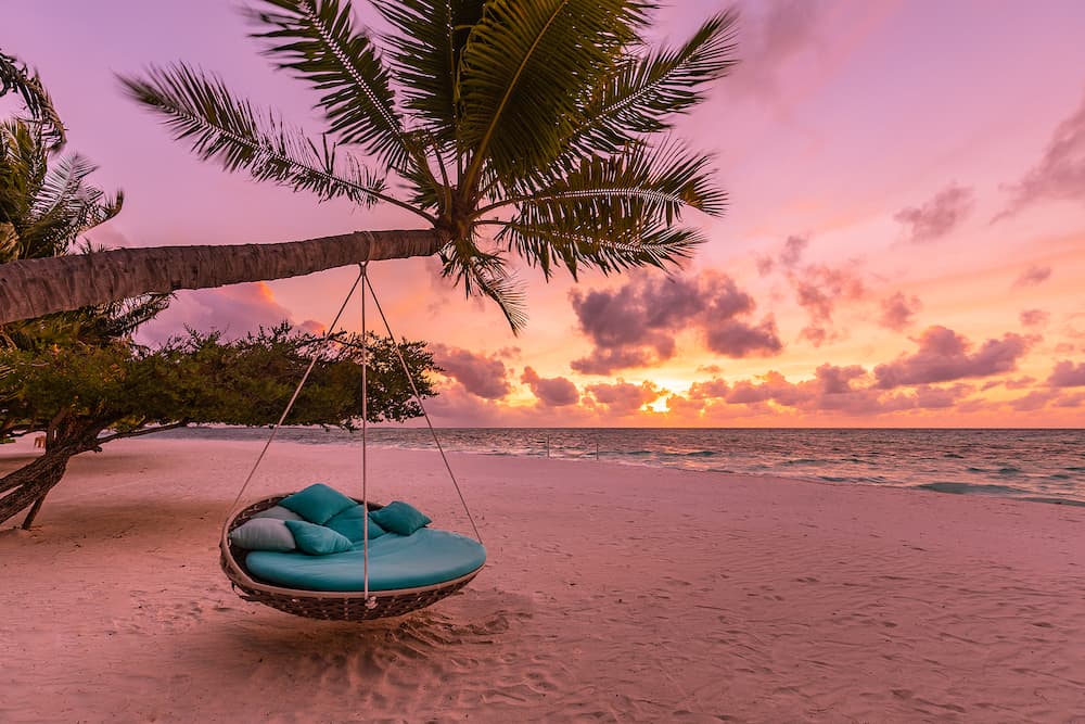 Tropical beach sunset landscape with beach swing or hammock hanging on palm tree with sunset sky white sand and calm sea for beach banner. Perfect beach scene vacation and summer holiday concept.