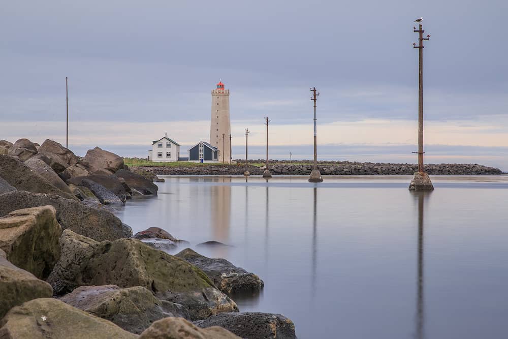 Lighthouse and uninhabited houses in the background. Lateral power pylons in the water during long-term exposure. Stone coastline of the island of Grotta near Reykjavik with clouds in the sky