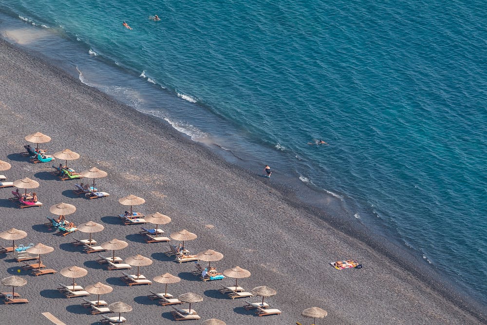 Kamari, Santorini Island, Greece- Black, Volcanic beach wit chairs lawn in Kamari resort. Coastline Aegean Sea.