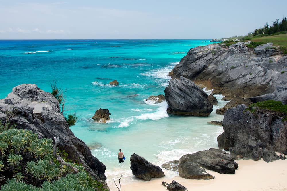 Scenic overhead landscape of man splashing in the waves of a beautiful, hidden Bermuda beach surrounded by stoney cliffs and jutting rocks and boulders.