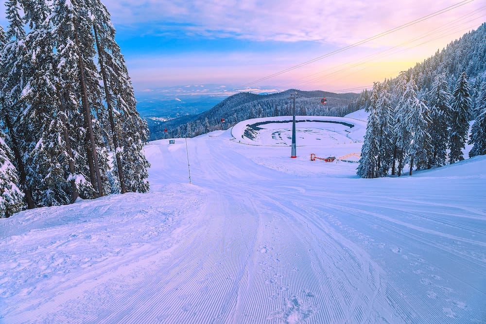 Fabulous snow covered pine trees and winter ski resort with colorful ski gondolas at sunrise. Ski slopes and frozen lake in Poiana Brasov resort, Transylvania, Romania, Europe