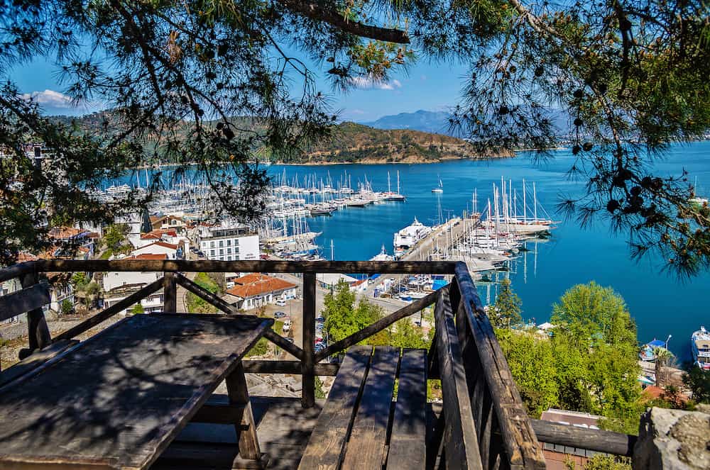 Fethiye, Turkey - View of Fethiye harbour with yachts and boats and mountains. There is a wooden table with benches for rest in the foreground. Mugla Province.