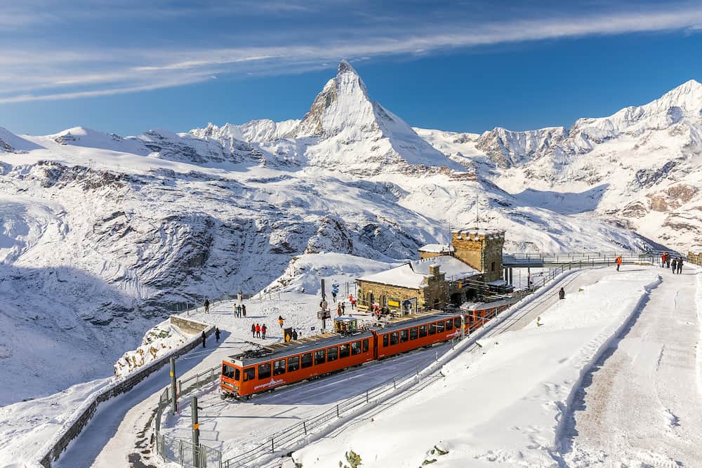 Gornergrat, Zermatt, Switzerland - Red cable car train on snowy railway at summit station with tourists and Matterhorn summit in winter.