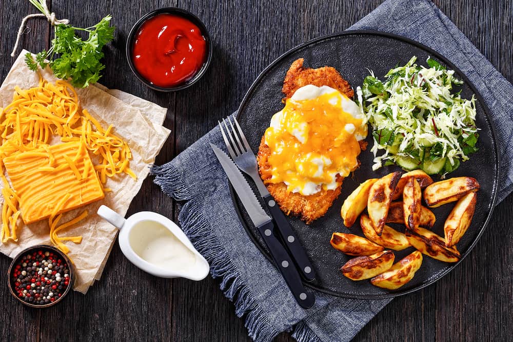 chicken parmo, chicken breast cutlet deep-fried in breadcrumbs, topped with creamy sauce and melted cheese, served with roasted potato and coleslaw on black plate with ingredients, flat lay