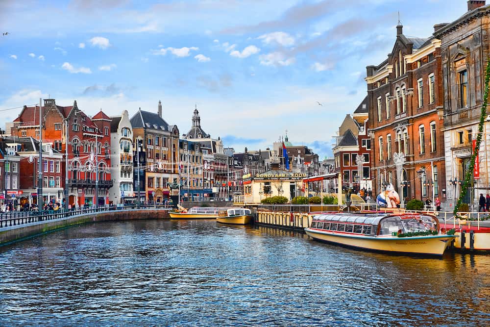 AMSTERDAM, NETHERLANDS - Picturesque views of the canal and city center of Amsterdam on a bright sunny day after Christmas. Canal tourist boats are moored to the embankment on background of beautiful old houses and a clear blue sky