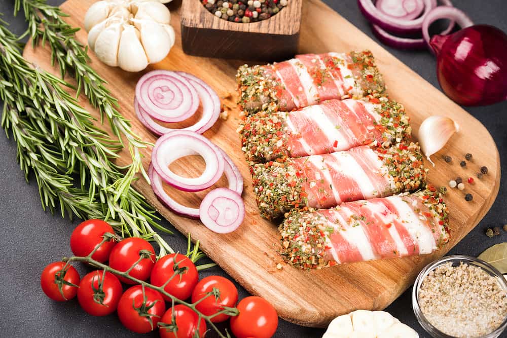 Balkan cevapcici or Romanian mititei. Traditional Balkan food. Raw minced meat rolls wrapped with bacon with seasonings and vegetables around over dark concrete background