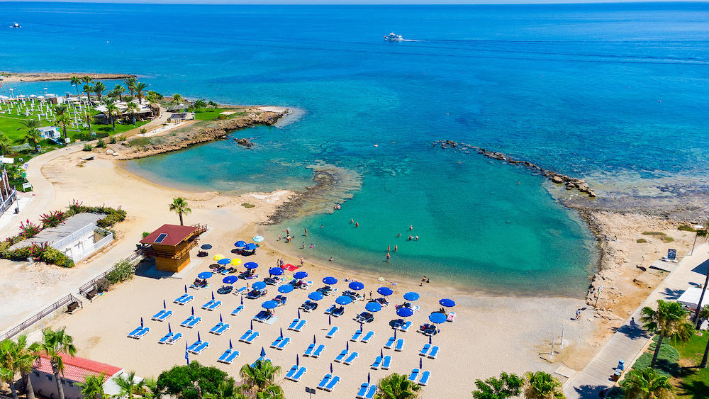 Aerial bird's eye view of Pernera beach in Protaras, Paralimni, Famagusta, Cyprus. The famous tourist attraction golden sandy bay with sunbeds, water sports, hotels, restaurants, people swimming in sea on summer holidays from above.