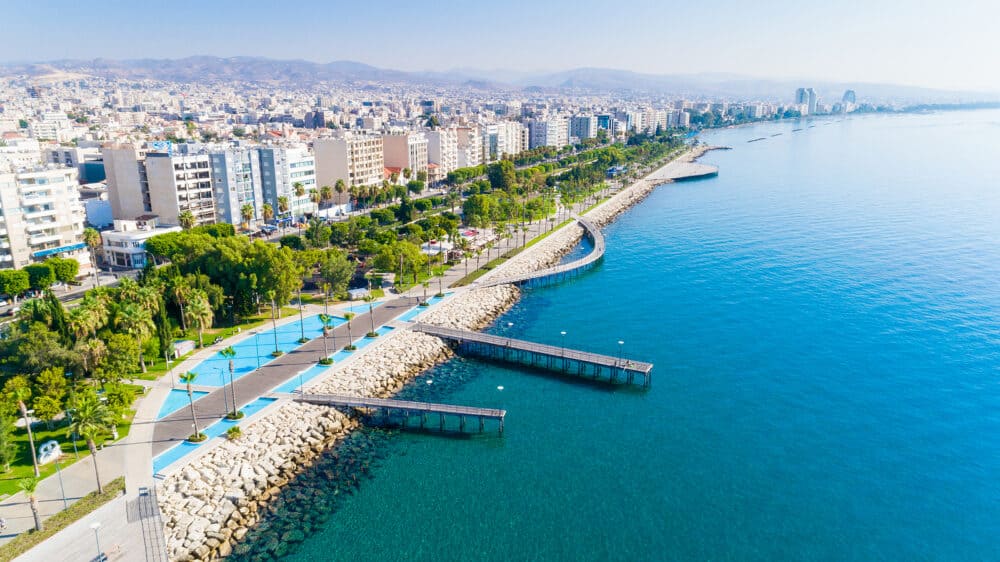 Aerial view of Molos Promenade park on the coast of Limassol city centre in Cyprus. Bird's eye view of the jetties, beachfront walk path, palm trees, Mediterranean sea, piers, rocks, urban skyline and port from above.
