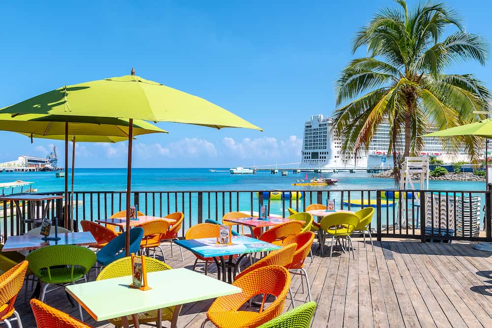 Ocho Rios, Jamaica - Tropical island Caribbean color chairs tables at Margaritaville Ocho Rios Restaurant and Sports Bar on the beach in Saint Ann. Norwegian Epic Cruise Ship in port.