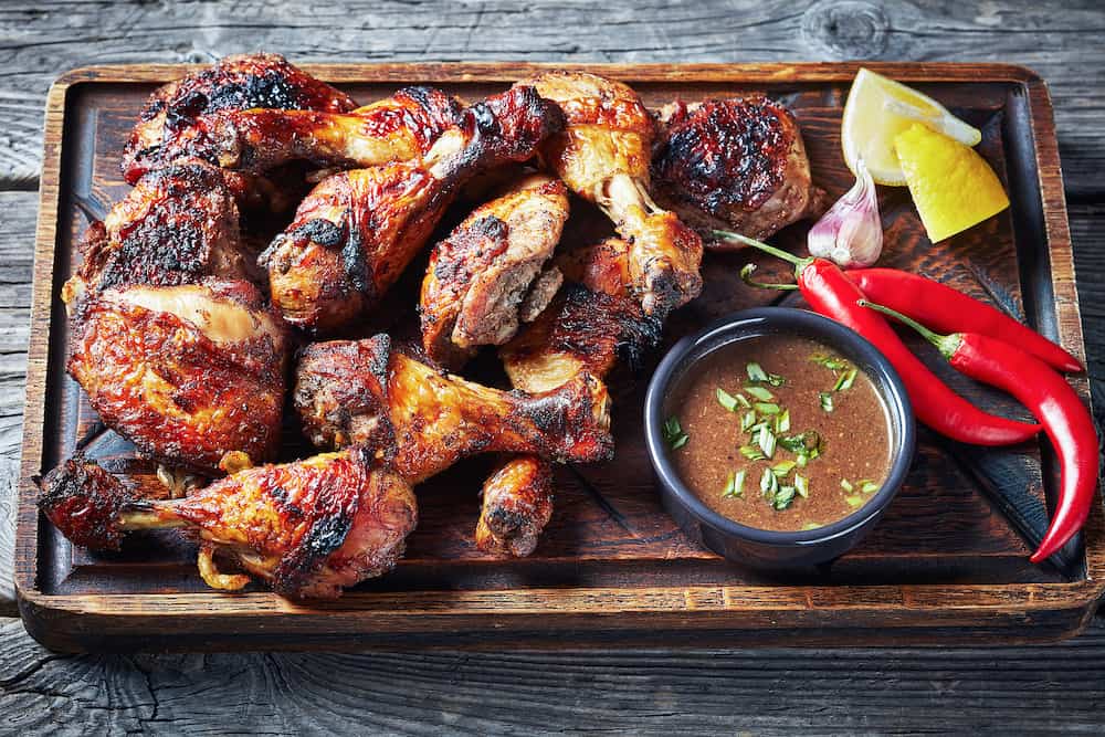hot Grilled Jamaican Jerk Chicken on a rude board with sauce and lemon on a rustic wooden table, horizontal view from above, close-up