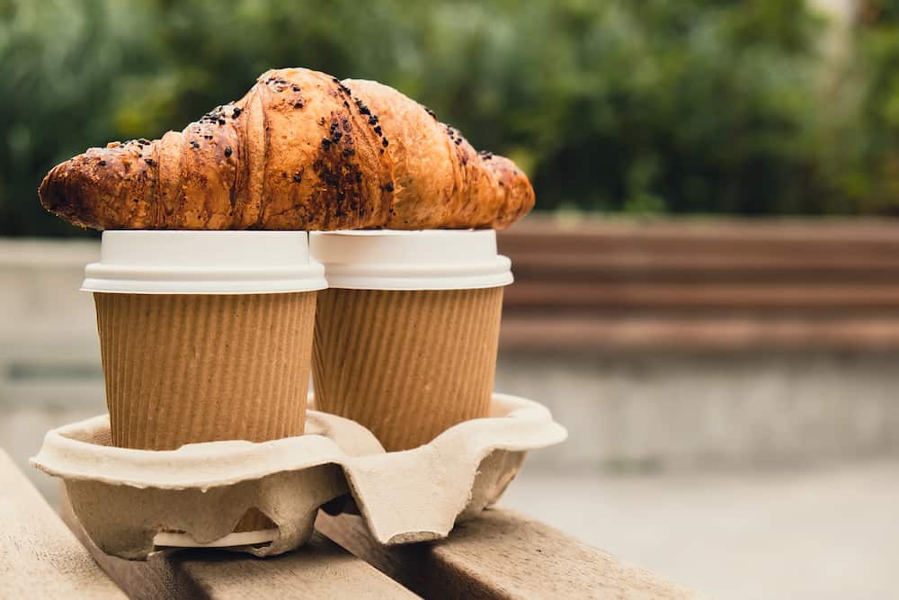 Fresh baked chocolate croissant on Two paper cups with lid for tea to go. Breakfast Coffee take away on the table. Take-out coffees with brown paper cup holder. Brown safety cardboard collars. Cardboard tray