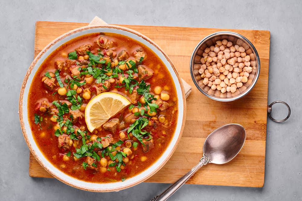 Moroccan Harira Soup in black bowl at grey concrete table top. Harira is Moroccan Cuisine dish with lamb or beef meat, chickpeas, lentils, tomatoes, ciliantro. Ramadan Iftar Food.