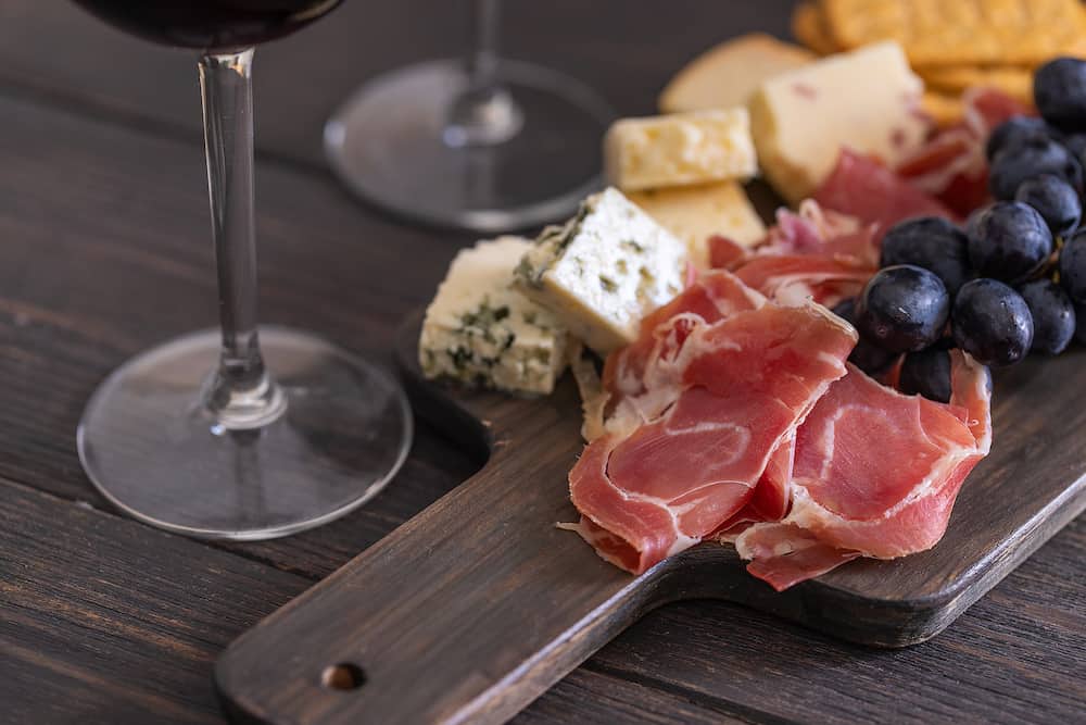 Wooden board with different snacks for red wine. Cheese assortment, ham, grapes and crackers. Wine snacks set background.