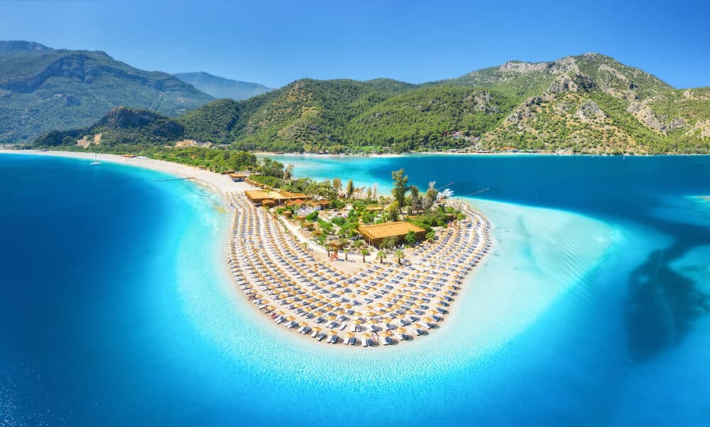 Aerial view of sea bay, sandy beach with umbrellas, trees, mountain at sunny day in summer. Blue lagoon in Oludeniz, Turkey. Tropical landscape with island, white sandy bank, blue water. Top view