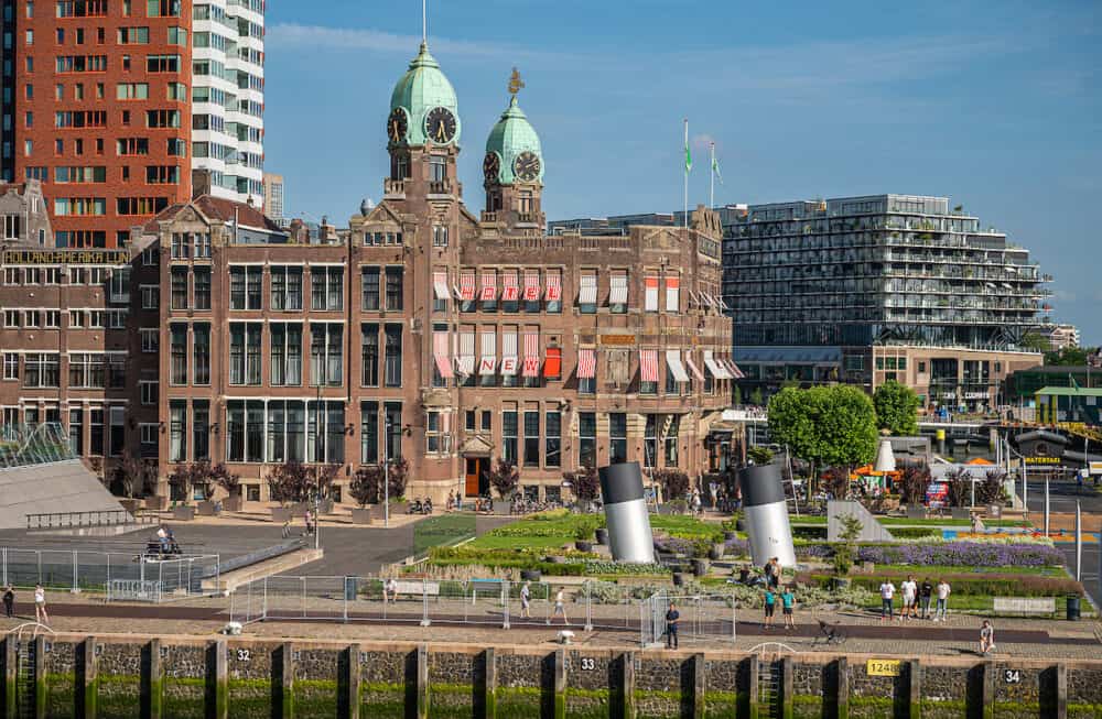 Rotterdam, Netherlands - Brown stone historic New York hotel with 2 green dome towers under blue sky on Kop van Zuid dock and quay. People present in garden. Other buildings in back