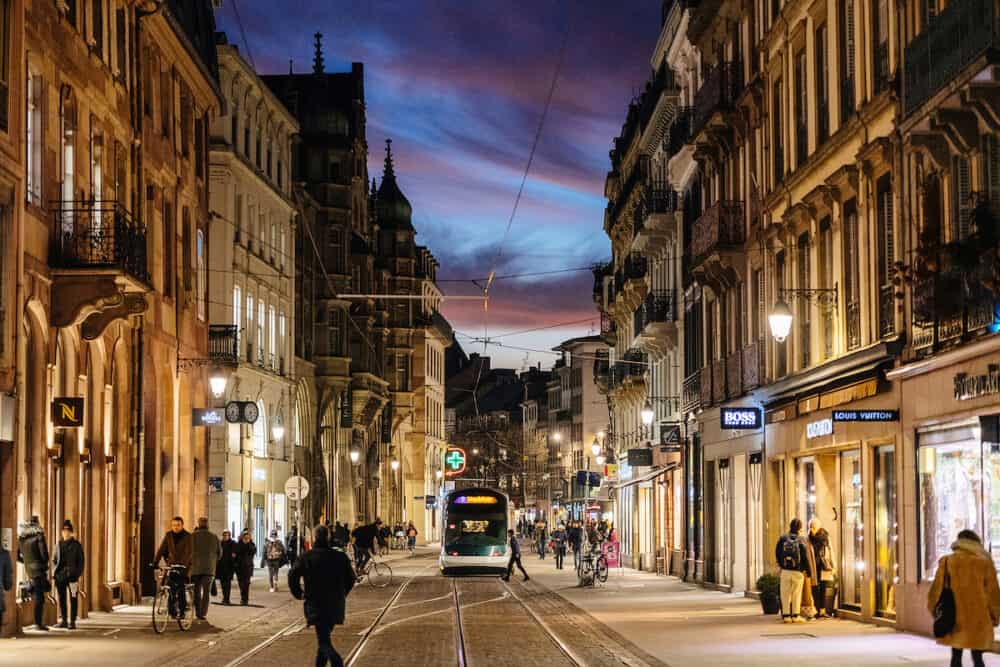 Strasbourg, France - People, tramways and open shops stores on the beautiful illuminated Rue de la Haute Montee street with beautiful clouds at dusk sunset