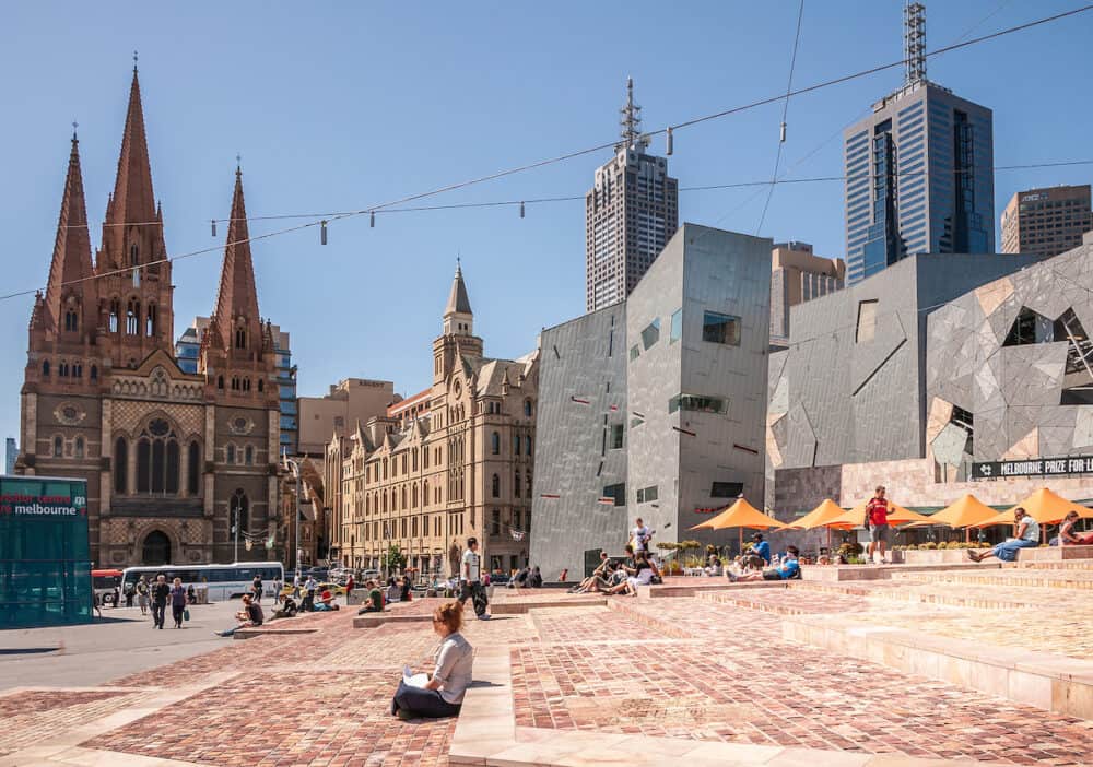 Melbourne, Australia - Federation square baking in sun under blue sky is surrounded by Saint Pauls Cathedral, Torrens University building and ACMI modern architecture building.