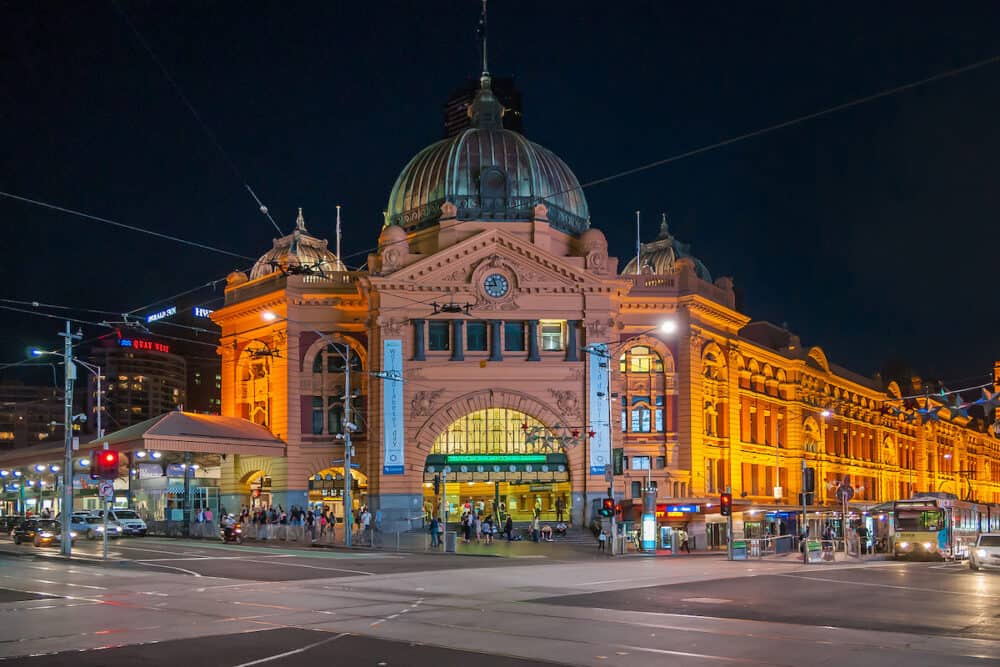 Melbourne, Australia - Lighted yellow historic building of Flinders Street Railway Station with its green-brown dome and flag at a dark night. Traffic of cars, trams and people.