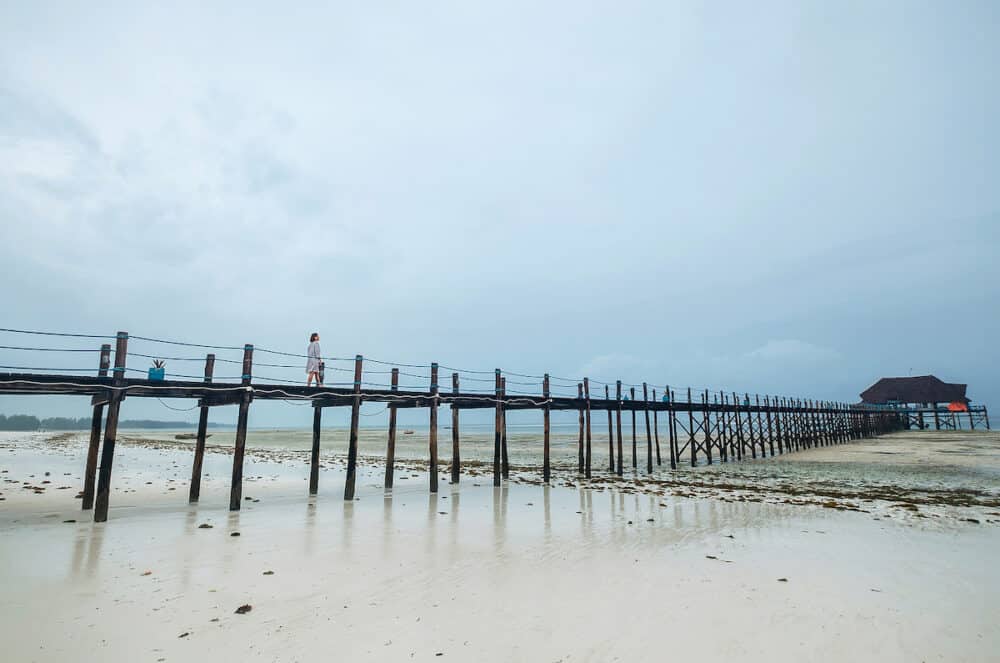 A woman dressed in light summer clothes walking on the Zanzibar island low tide sandy beach wooden pier. Careless vacation in the tropical countries concept image. Kiwengwa, Tanzania.